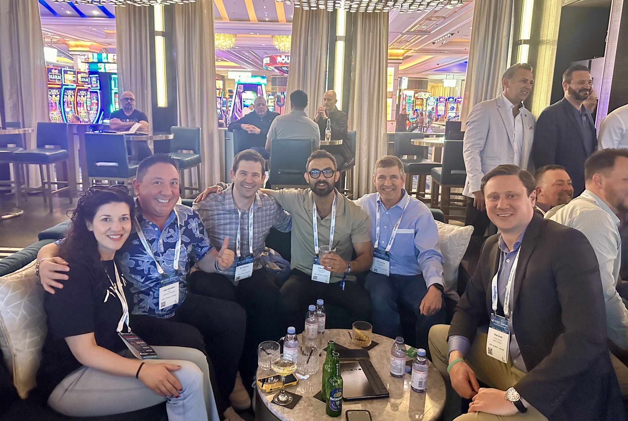 A group of six Channel Partners, wearing conference badges, sit smiling around a table with drinks in a lively casino lounge, with slot machines and other people in the background.